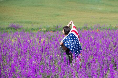 Attractive woman holding Flag of the United States in beautiful summer field on a clear, sunny day. Celebrating Independence Day, National holiday concept.の写真素材