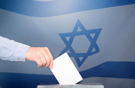 Election in Israel. Hand of voter putting vote in the ballot box. Waved Israel flag on background.の写真素材