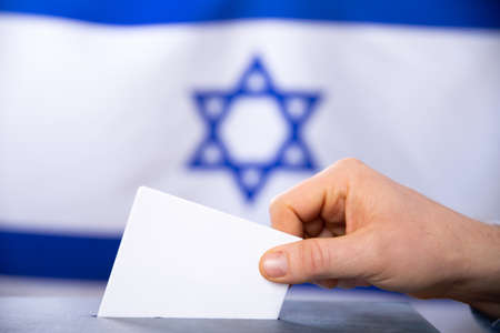 Election in Israel. Hand of voter putting vote in the ballot box. Waved Israel flag on background.の写真素材