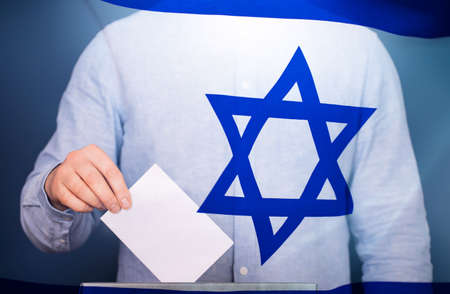 Election in Israel. Hand of voter putting vote in the ballot box. Waved Israel flag on background.の写真素材