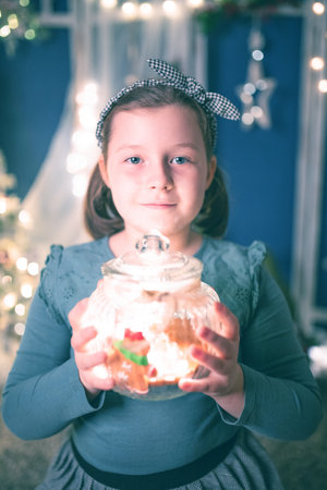 Young girl holding a candy jar with homemade gingerbread cookies.Christmas lights on backgroundの写真素材