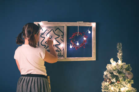 Cute little girl writing on snowy window, Christmas decorations and waiting for Santa