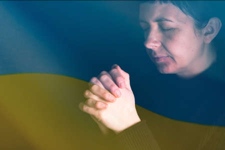 Sad woman with Ukraine flag at background praying. Concept of standing with Ukrainian nation in war with Russia.の写真素材