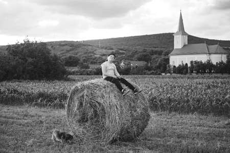 Caucasian girl having fun sitting on top of a golden hay bale on summer field near farm. Happy childhood and freedom concept. Rural countryside scenic landscapeの写真素材