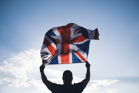 The flag of England waving in the hands of a man against the blue sky. View from the back. Great Britain, victory and successの写真素材
