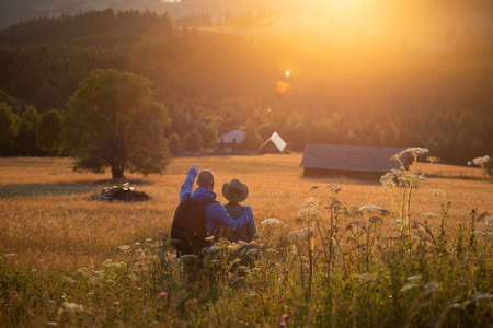 Hiker couple in summer meadow looking at magic sunset over hills. Relaxation, nature and tourism concept.の写真素材