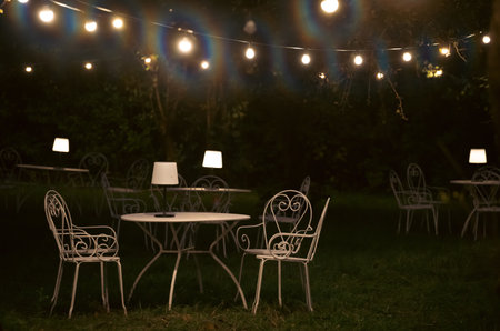 Victorian style cafe with white empty metal chairs and tables in outdoor at evening. Wrought iron furniture, old fashion cozy cafe terrace, restaurant tables waiting for customers at an outdoor terrace.の写真素材