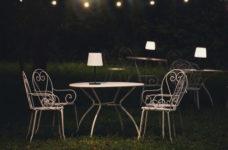 Victorian style cafe with white empty metal chairs and tables in outdoor at evening. Wrought iron furniture, old fashion cozy cafe terrace, restaurant tables waiting for customers at an outdoor terrace.の写真素材