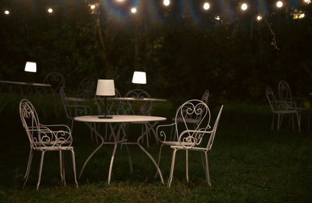Victorian style cafe with white empty metal chairs and tables in outdoor at evening. Wrought iron furniture, old fashion cozy cafe terrace, restaurant tables waiting for customers at an outdoor terrace.の写真素材