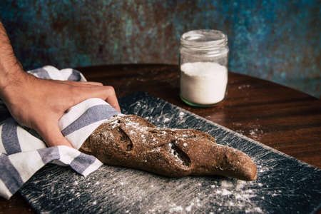 A man hand holding a handmade rye bread wrapped with a cloth over a stone table and a flour jar next...