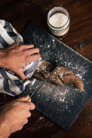 A man hand is holding and slicing a handmade rye bread wrapped with a cloth over a stone table, there is a flour jar next to it.の写真素材