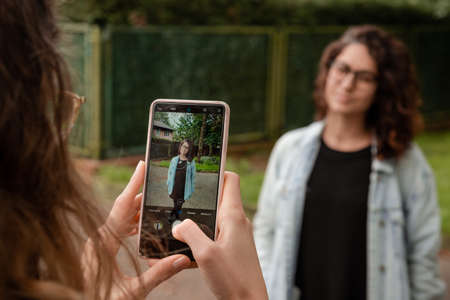 Young woman taking a picture of her friend with a mobile phone in the park.の写真素材