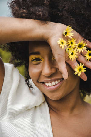 An african american young woman smiling with yellow makeup and yellow flowers in her hands outdoors.の写真素材