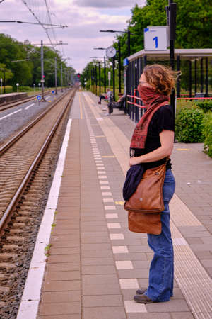 Young caucasian woman wears an scarf as a face mask while waiting on a trainstation, protecting herself and others from coronavirus.のeditorial素材