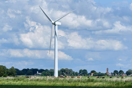 Typical Dutch farmland landscape with dike and wind turbine with cows in pasture and village in background on a warm windy day. the Netherlandsの写真素材