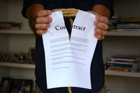 Tied hands tearing contract paper in front of bookcase. Bound man hands rip paper contract, breach of contractの写真素材