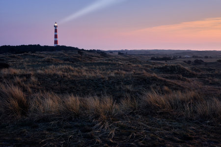 Historic lighthouse of Hollum with light beam, Ameland with a red and orange sky during sunset, sunrise. Dark dunes with stern grass in the foreground.の写真素材