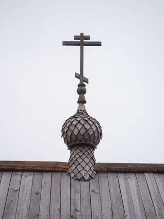 Picture of the Cross on the top of a wooden dome. The Cross on the wooden dome against the background of the cloudy sky. Roof of the ancient church made of brown wooden planks.の写真素材