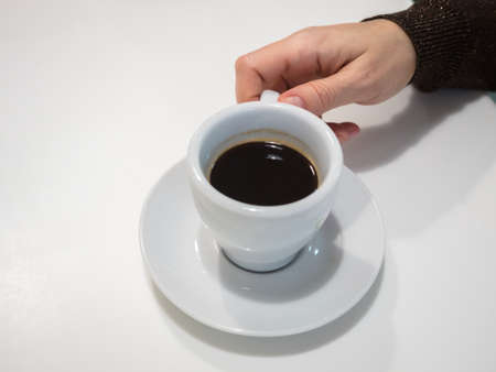 Top view of humans hand with cup of coffe standing on a saucer on white background. White cup with...