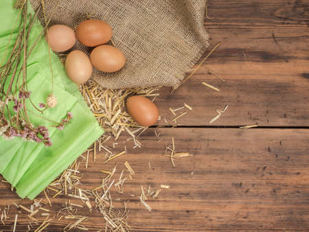 Rural creative background with brown eggs, burlap, straw, green paper and dry flowers on wooden table from old planks. Vintage, rustic background for Easter postcards, restaurant menus or advertisingの写真素材