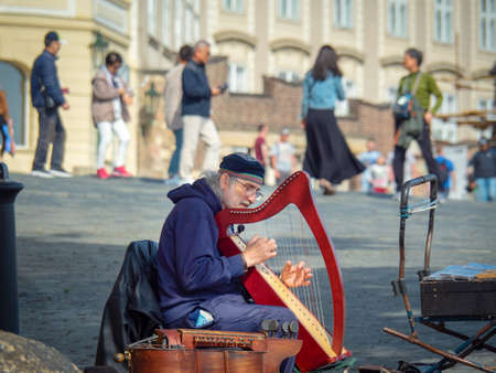 Prague, Czech republic - april 2019: Older man sitting on Charles bridge and playing on harp. Street musical player with his harp. Tourists walking on bridge in Prague, blurred backgroundのeditorial素材