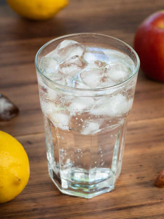 Close up view on glass full of water and melting ice cubes. Yellow lemons, red apple and glass on...