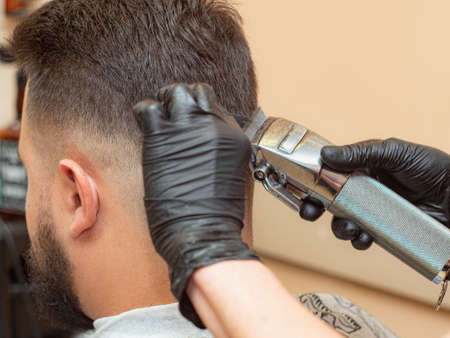Male in barbershop, making modern hairdo, close up view. Stylist cutting hair with clipper. Hands in black rubber gloves. Beige interior of barbershop. Selective soft focus. Blurred backgroundの写真素材