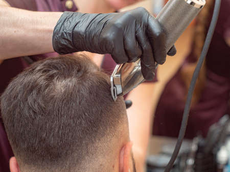 Females hand in black rubber glove cutting hair with clipper, close up view. Stylist making modern...