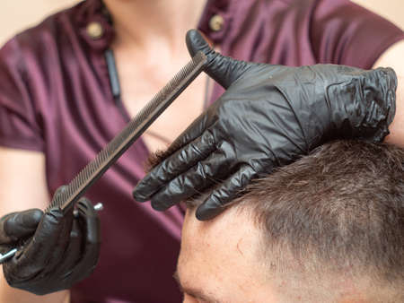 Female making modern hairdo for client, close up view. Stylists hands in black rubber gloves cutting hair with comb and scissors. Selective soft focus. Blurred backgroundの写真素材
