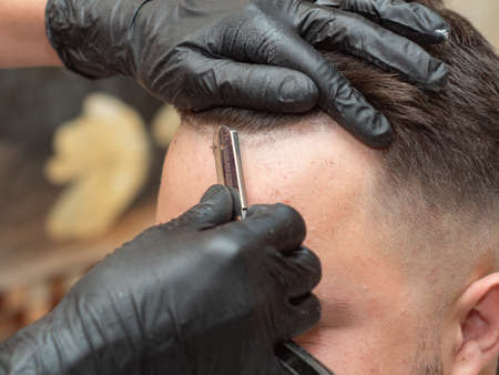 Stylist shaving clients forehead with open razor in barbershop, close up view. Hands in black rubber...