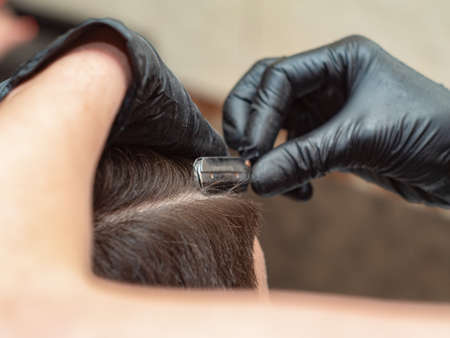 Hairdressing stylist cutting hair with straight razor, close up view. Females hands in black rubber gloves with open razor. Photographed in barbershop. Selective soft focus. Blurred backgroundの写真素材