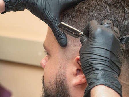 Stylist shaving males temple with open razor, close up view. Masters hands in black rubber gloves. Photographed in barbershop. Selective soft focus. Blurred backgroundの写真素材
