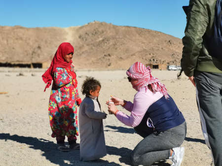 Bedouin village in desert, Egypt - February 2020: tourists and bedouins kids, middle view. Woman gives pack of juice to small boy. Selective soft focus. Blurred backgroundのeditorial素材