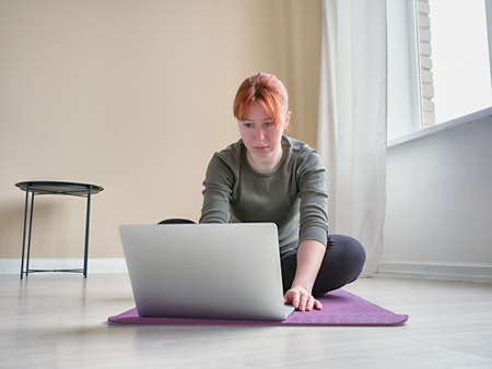 Front view to woman typing on laptop. Girl sitting on yoga mat near window. Concept of lifestyle in self-isolation. Online training and meditationの写真素材