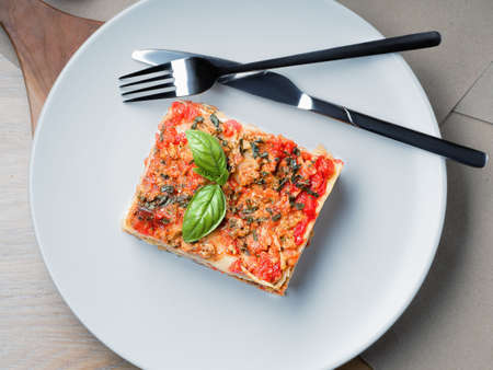 Piece of lasagna. White plate with fork and knife. Basil leafs, tomatoes and zucchini on table. Menu...