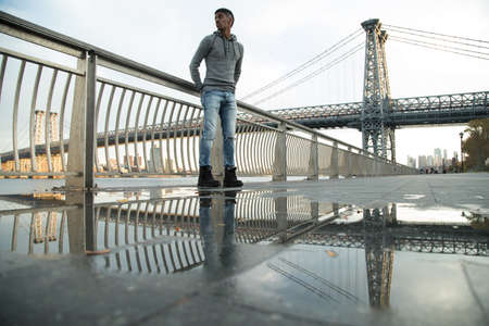 A young, black man walks along New York City's East River with Brooklyn's Williamsburg Bridge in the background. Shot during the Autumn of 2016.の写真素材