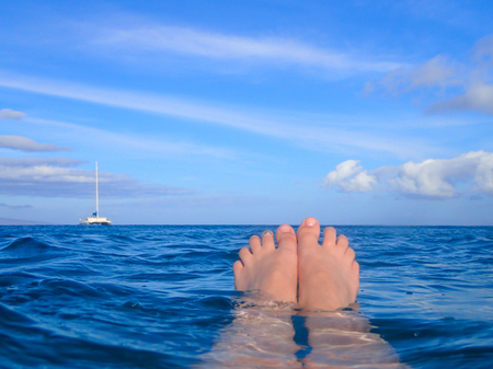 Toes emerge from ocean as person floats on surface with boat on horizon under blue skyの写真素材