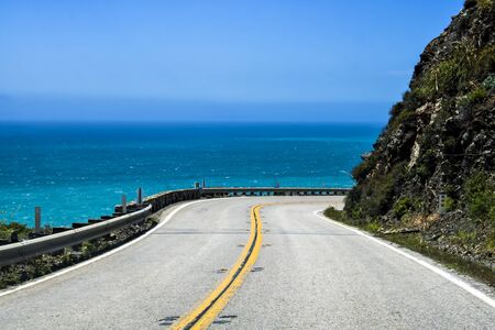 Highway 1 in California curved around a cliff edge with blue ocean in background.