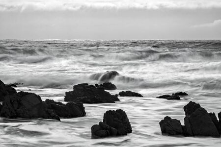 California black and white ocean seascape in motion with waves breaking over black rocks under glowing sky.の写真素材