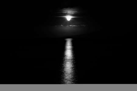 Black and white image of full moon dripping beneath clouds as it sets over the ocean with light reflecting on the sea and moon resembling alien spacecraft.の写真素材