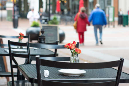 A couple walk hand in hand along an outdoor mall. In the foreground is a black metal table with a simple arrangement of roses with a white napkin and plates.の写真素材
