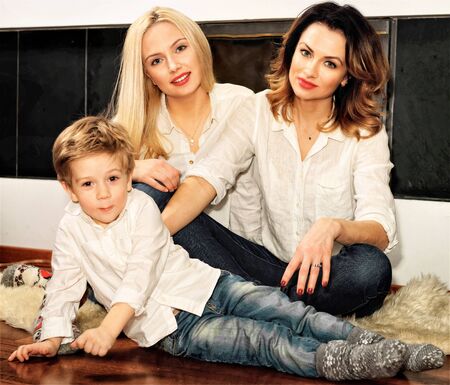 Beautiful family consisting of mother with ginger hair, daughter with blond hair and a son sitting on the fur carpet at the fireplace. Fire in the fireplace is burning.の写真素材
