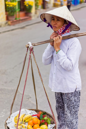 Hoi An, Vietnam - February 16, 2016: Asian trader carrying fresh fruit in bowls on her shoulders in the street in Hoi An, Vietnam. Rambutan, mango, banana and mandarin.のeditorial素材