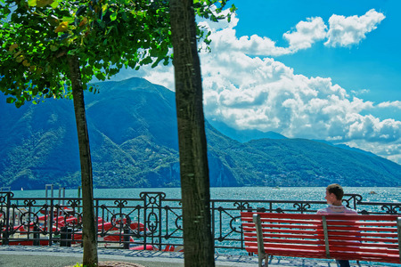 Man sitting on the bench at the promenade of the luxurious resort in Lugano on Lake Lugano and Alps mountains in Ticino canton of Switzerland.の写真素材