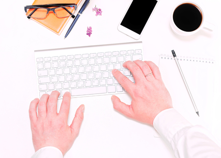 Man working with computer keyboard and workplace with mobile phone, cup of coffee, notebook, pen, pencil, glasses and lilac flowers on the white table background. Flat lay, top viewの写真素材