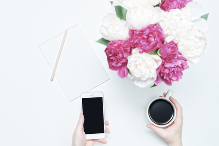Working place with hands holding cup of coffee and mobile phone, paper, pen and white and pink peony flowers on white table background. Flat lay, top viewの写真素材