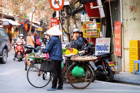 Hanoi, Vietnam - February 21, 2016: Woman wears traditional hat selling fruit from bicycle in street of Hanoi in Vietnam. Street market vendor with Vietnamese cousine. Poor merchant. Food seller.のeditorial素材
