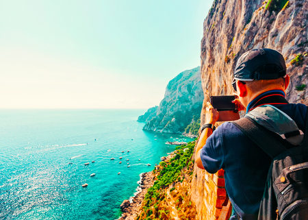 Man taking photos of Marina Piccola in Capri Island in Italy at Naples. Landscape with Mediterranean Sea at Italian coast. Anacapri in Europe. View on Faraglioni. Amalfi scenery and Solaro mountainの写真素材