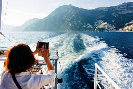 Tourist taking photos on camera during cruise excursion tour on boat in Tyrrhenian Sea in Positano town of Amalfi Coast, Italy, summer. Beautiful Mediterranean view near Sorrento. Travel and tourismの写真素材