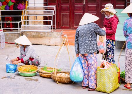 Hanoi, Vietnam - February 17, 2016: Women merchants in traditional Vietnamese hats selling and buying food on street market in Hoi An in Vietnam in Asia. Local small business with sellers and vendorsのeditorial素材